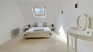 Une chambre à coucher avec un lit, une table de chevet avec une lampe, une commode avec un miroir, un radiateur et une fenêtre donnant sur des arbres.