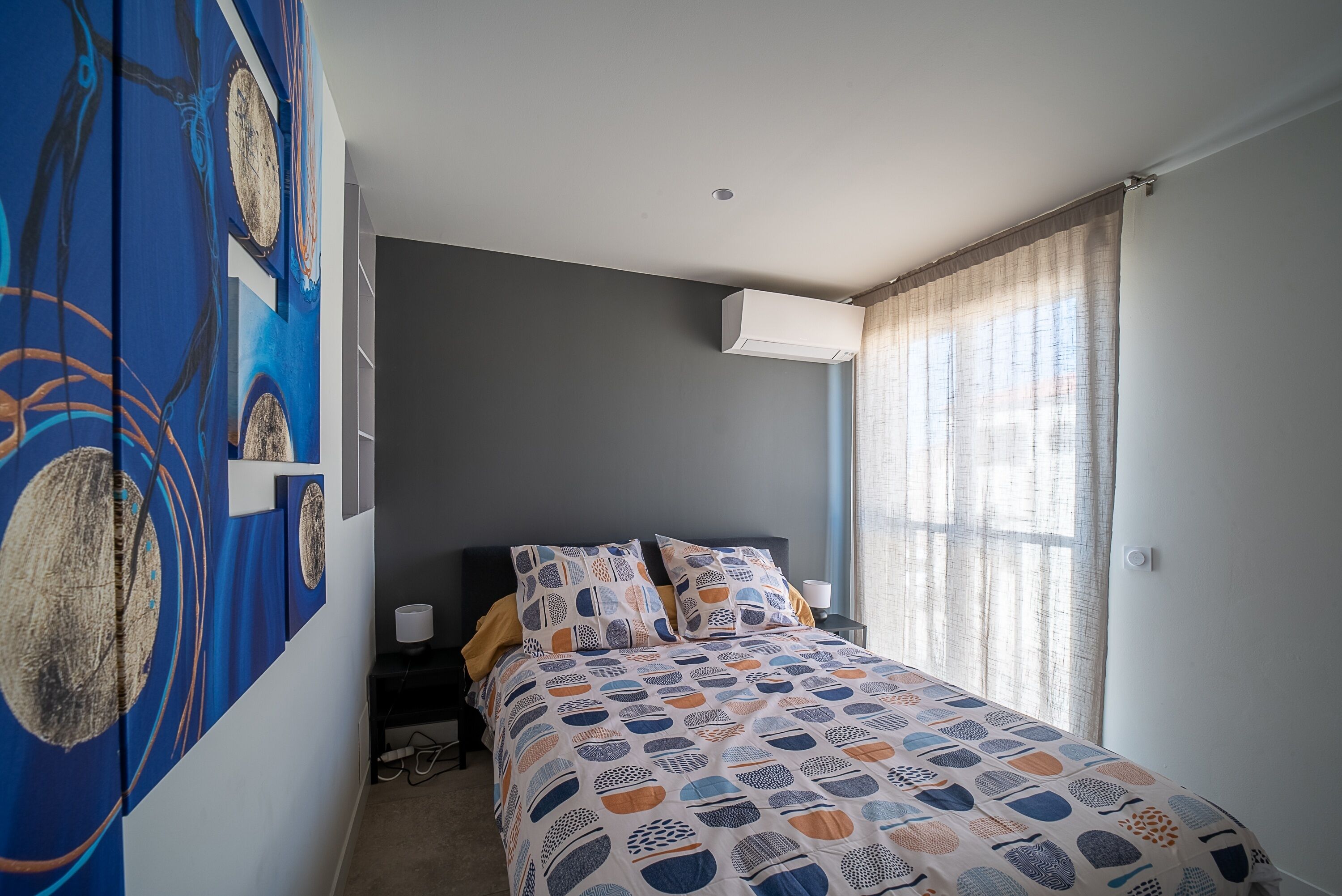 A bedroom with a bed, a nightstand, a wall-mounted air conditioner, and a window with curtains.