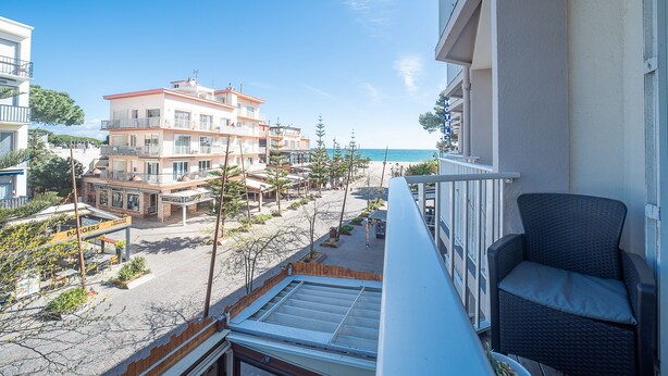 Un balcón con una silla de mimbre, con vista a una playa que incluye edificios y un restaurante.