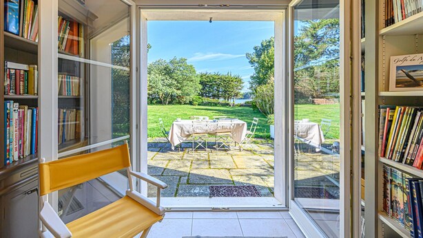 A room with a large glass door leading to an outdoor area with a table and chairs, surrounded by bookshelves filled with books.