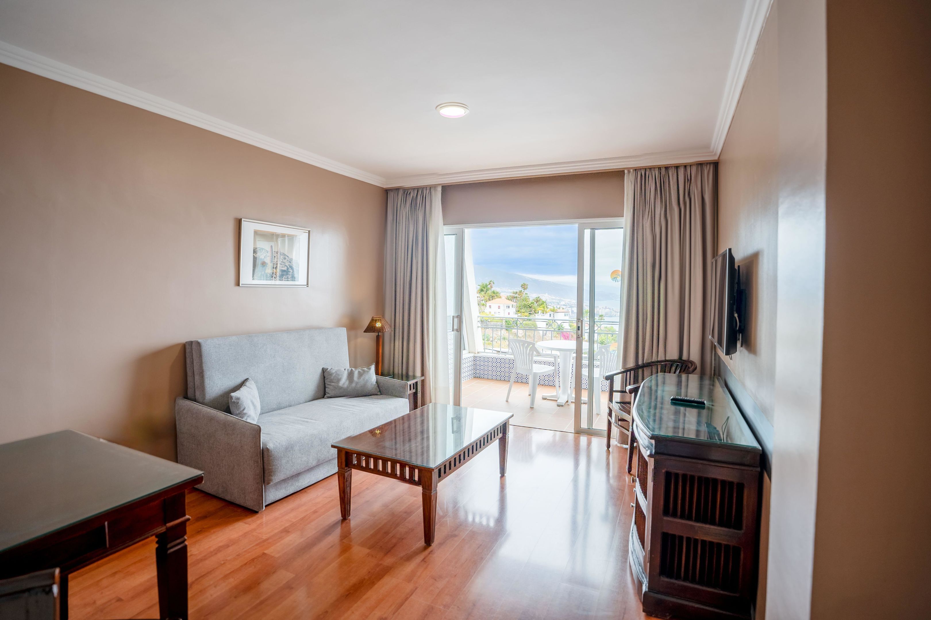 A modern living room with a sofa, a glass-top coffee table, a TV mounted on the wall, and a balcony with a view.