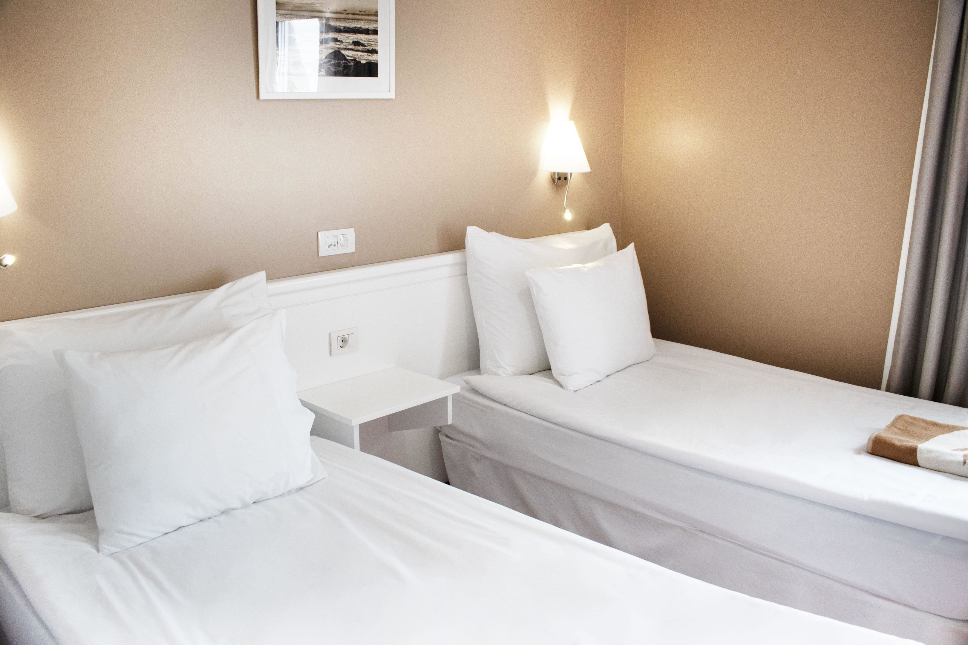 A hotel room with two beds, a nightstand, a wall-mounted lamp, and a framed picture on the wall.