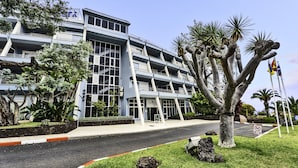 Un hôtel à plusieurs étages doté d’une architecture moderne, avec des balcons et un jardin bien entretenu où poussent des plantes tropicales.