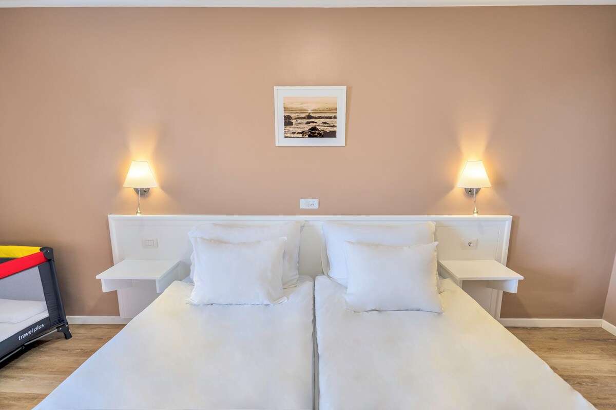 Un lit double avec du linge de lit blanc, deux tables de chevet avec des lampes, un tableau encadré au mur et une petite table sur laquelle se trouve un objet rouge et jaune.