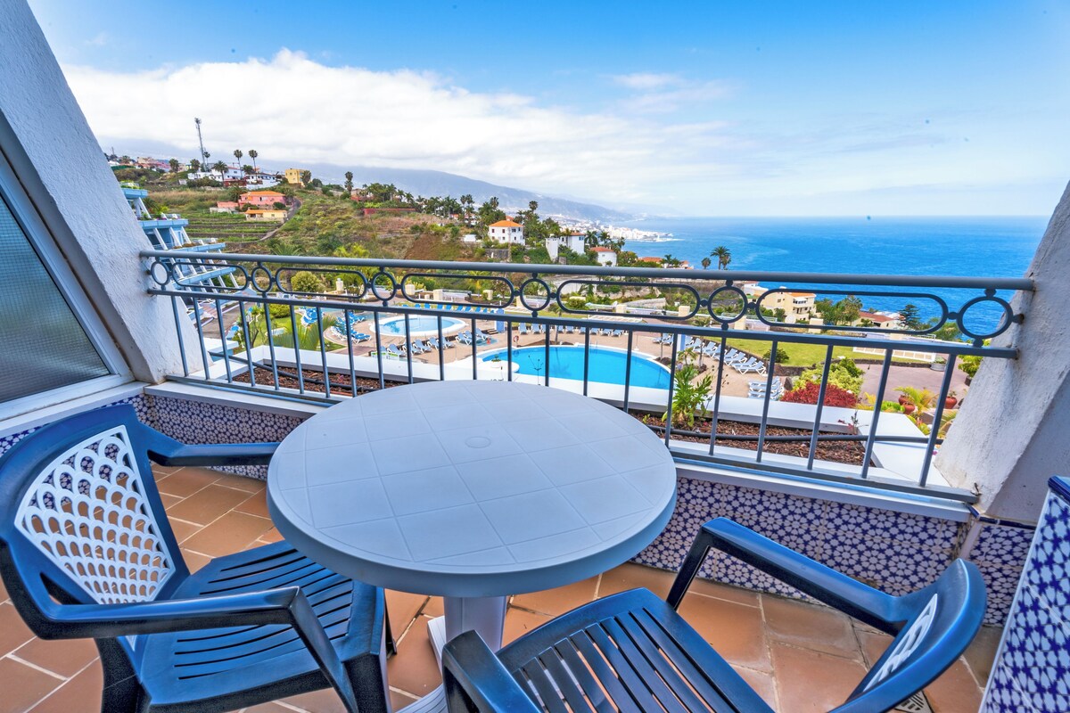 Ein Balkon mit rundem Tisch und Stühlen, der Blick fällt auf einen Pool und die Küste.