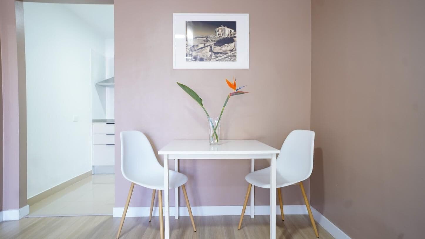 A dining area with a white table and two white chairs, a vase with flowers, and a framed picture on the wall.