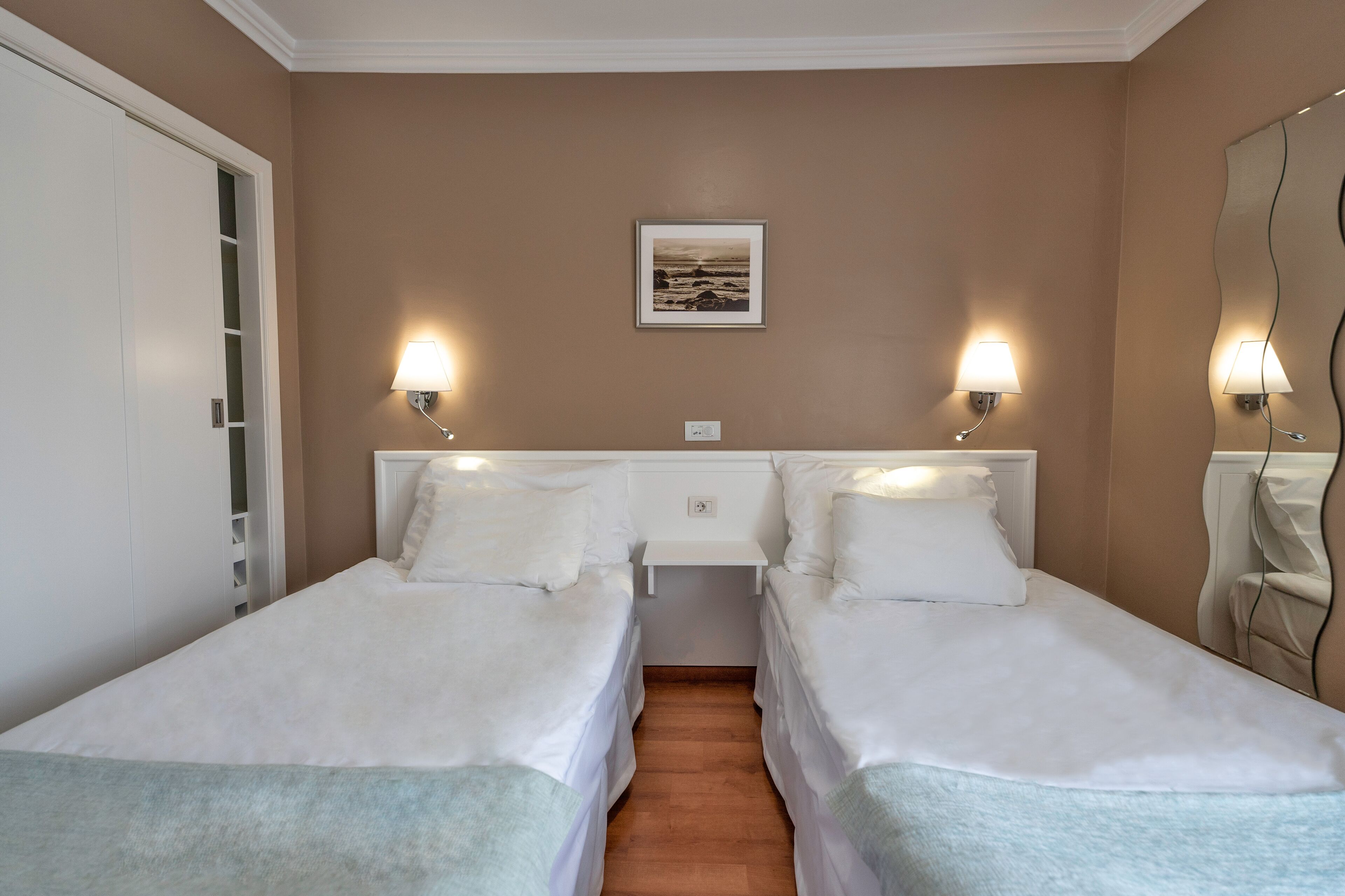 Een kamer met twee bedden, een witte nachtkastje, een lijst met een afbeelding aan de muur, en wandlampen.
