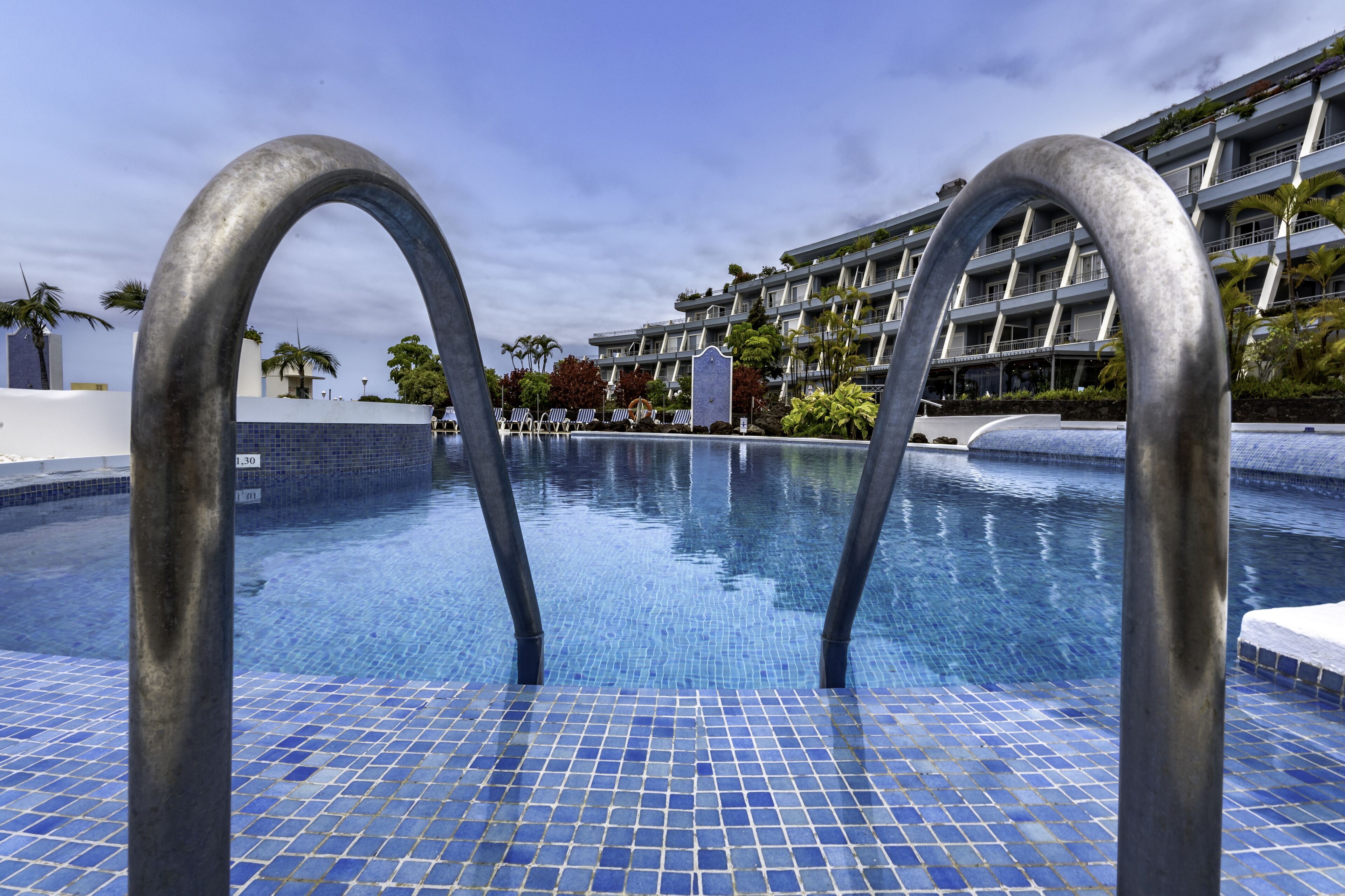 Uma piscina com azulejos azuis em mosaico, rodeada por um edifício moderno com varandas e palmeiras.