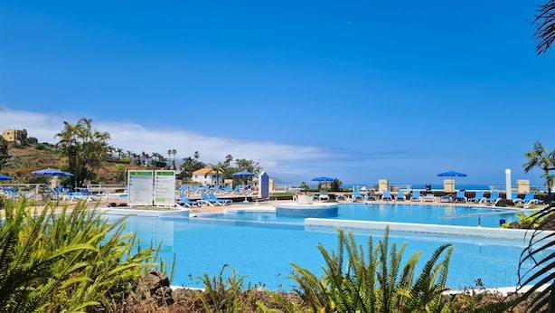 Un espace piscine doté de nombreuses chaises longues et de parasols, entouré de palmiers et surplombé d’un ciel d’un bleu limpide.