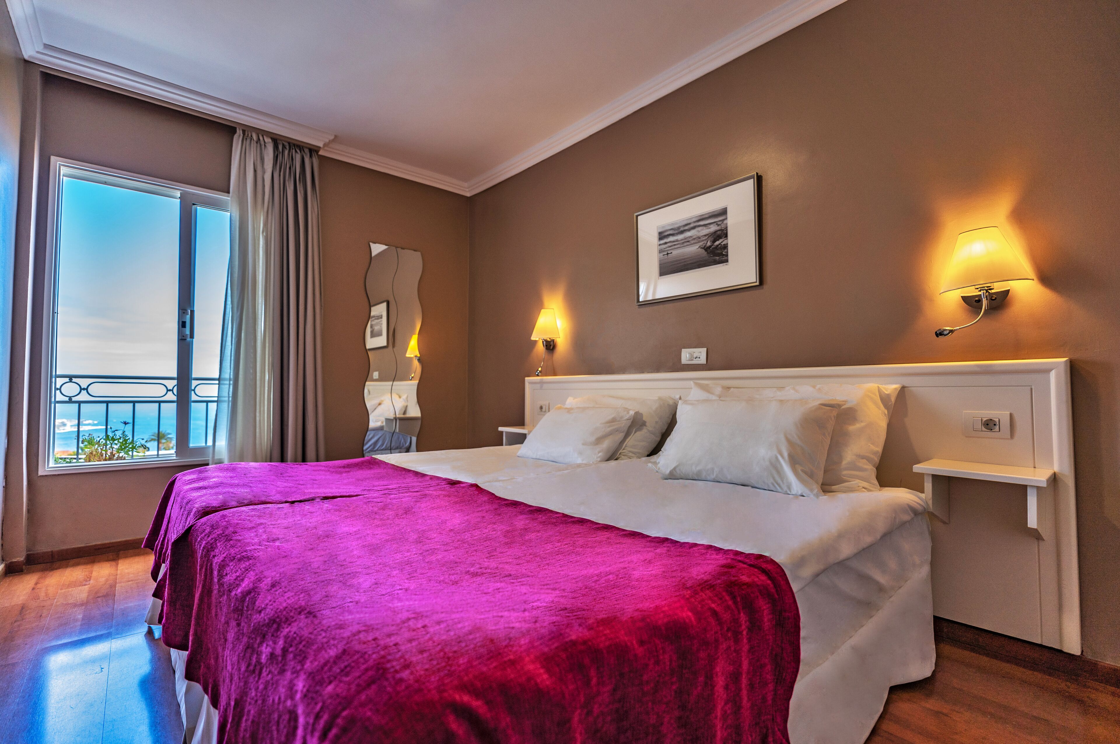 A hotel room with a large bed, two bedside lamps, a window with a view, and a framed picture on the wall.