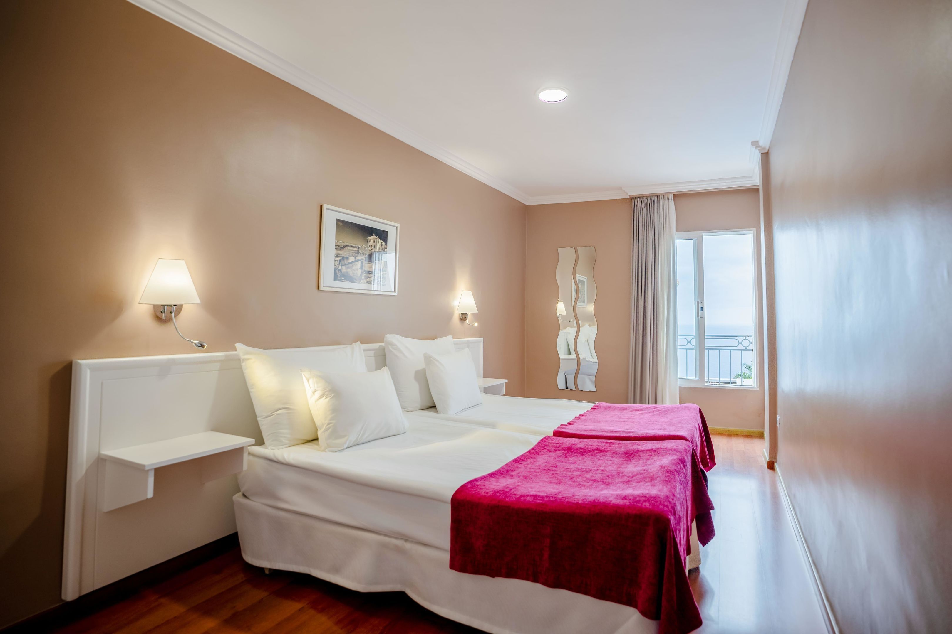 A hotel room with a large bed, bedside tables, a window with curtains, and a view of the outdoors.