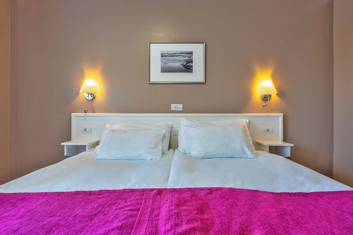 A hotel room with a large bed, two bedside lamps, a framed picture on the wall, and a white nightstand.