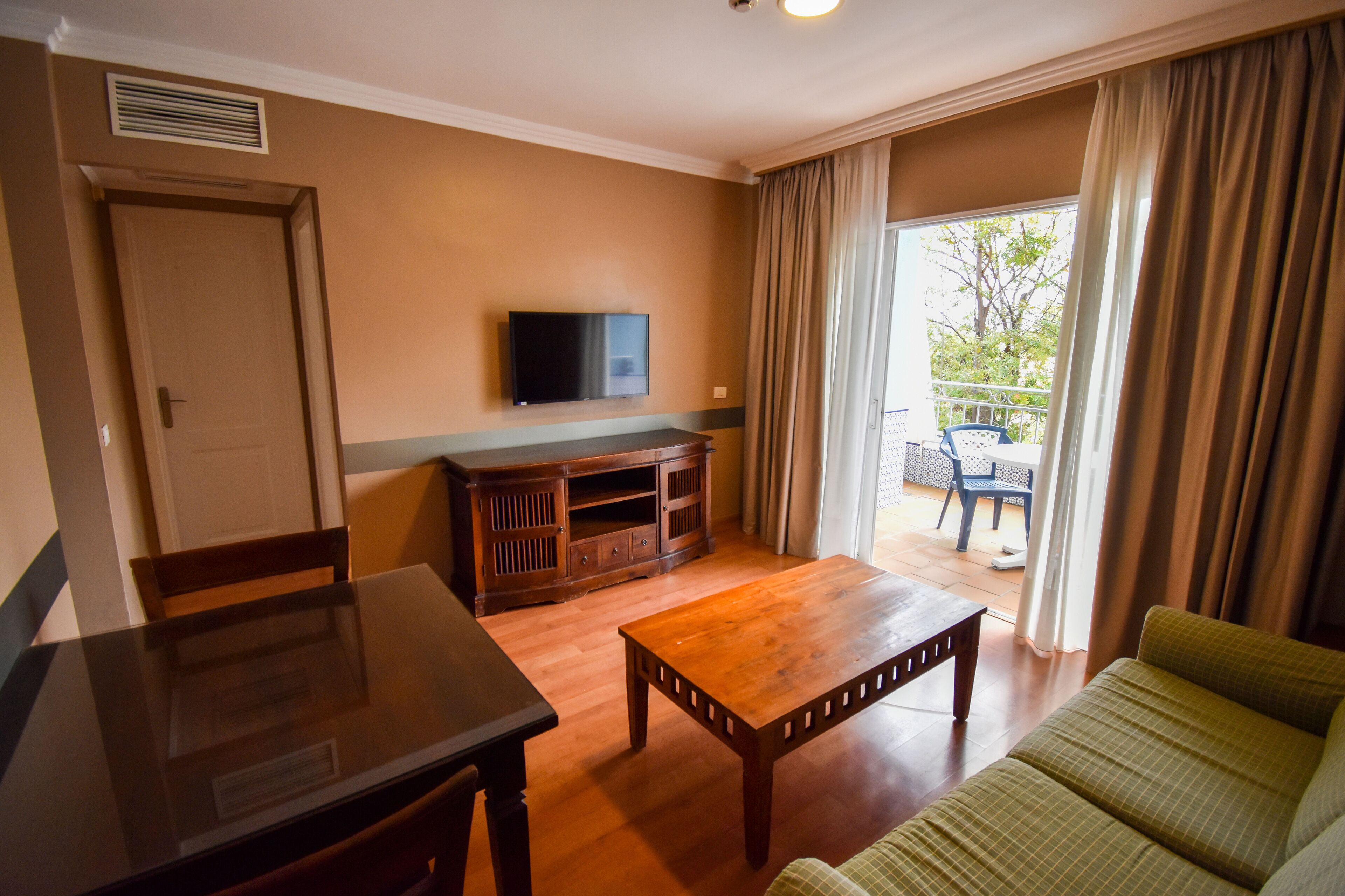 A living room with a wooden table, a sofa, a TV mounted on the wall, and a balcony with a chair.