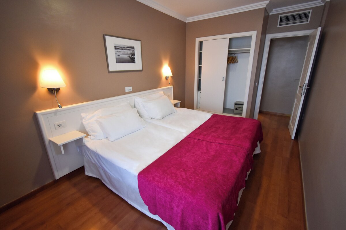 Un lit bien fait, avec du linge de lit blanc et une couverture rose, deux lampes de chevet, un tableau encadré au mur et une armoire avec un miroir.