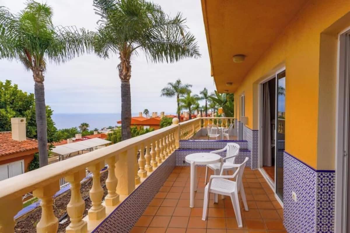 Un balcon doté d’un sol carrelé, de chaises blanches et offrant une vue sur des palmiers et des immeubles.
