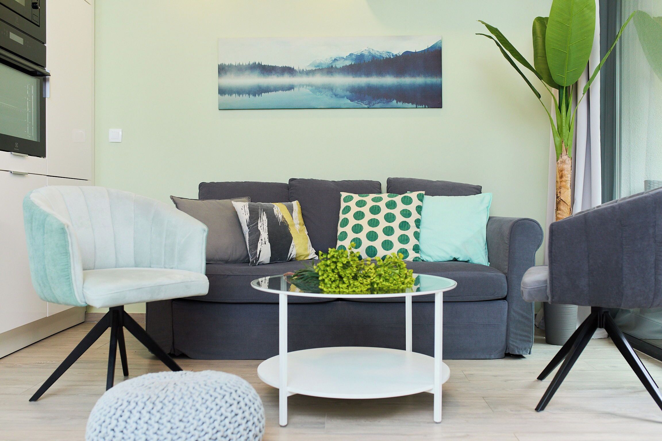 Un salon moderne avec un canapé gris, un fauteuil bleu clair, une table basse blanche ornée d’une plante verte et une grande fenêtre donnant sur un paysage montagneux.