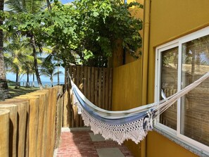 A hammock tied between a wooden fence and a yellow wall, with a view of palm trees and the ocean.