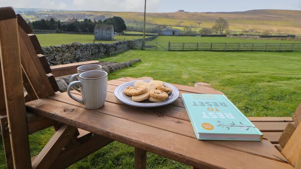 Una panca di legno con un piatto di biscotti, due tazze e un libro intitolato “REST TO REGENERATE” in un ambiente naturale suggestivo.