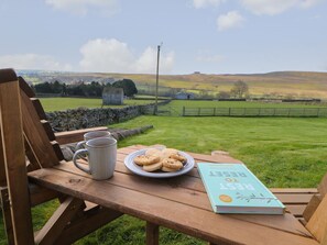 A wooden bench with a plate of cookies, two mugs, and a book titled "REST TO REGENERATE" in a scenic outdoor setting.