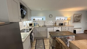 A modern kitchen with white cabinets, a built-in microwave, a dishwasher, and a stone accent wall. - Welcoming 2 Bed Charming Self Catering Cottage (Cheddar)