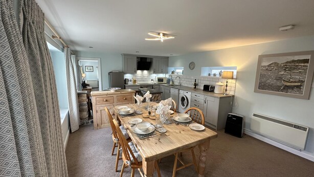 A compact kitchen with a dining area, featuring a wooden table set for four, a washing machine, and a microwave. - Welcoming 2 Bed Charming Self Catering Cottage (Cheddar)