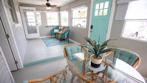 A room with a glass table surrounded by wicker chairs and a potted plant. There is a door with a window, a blue chair, and a small table with a vase.