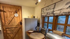 A compact kitchen with a wooden door, a window with curtains, a toaster, a kettle, and a bread box. - Studio Annex (Cambridge)
