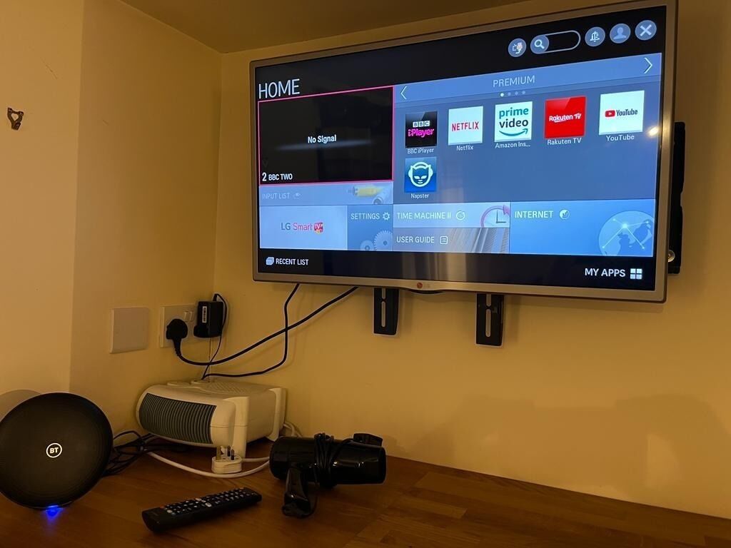 A television mounted on a wall with a remote control, a speaker, and a power strip on a wooden table.
