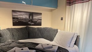 A modern bedroom with a large bed, a dark grey blanket, and a folded towel. A framed picture of a bridge is on the wall. - Studio Annex (Cambridge)