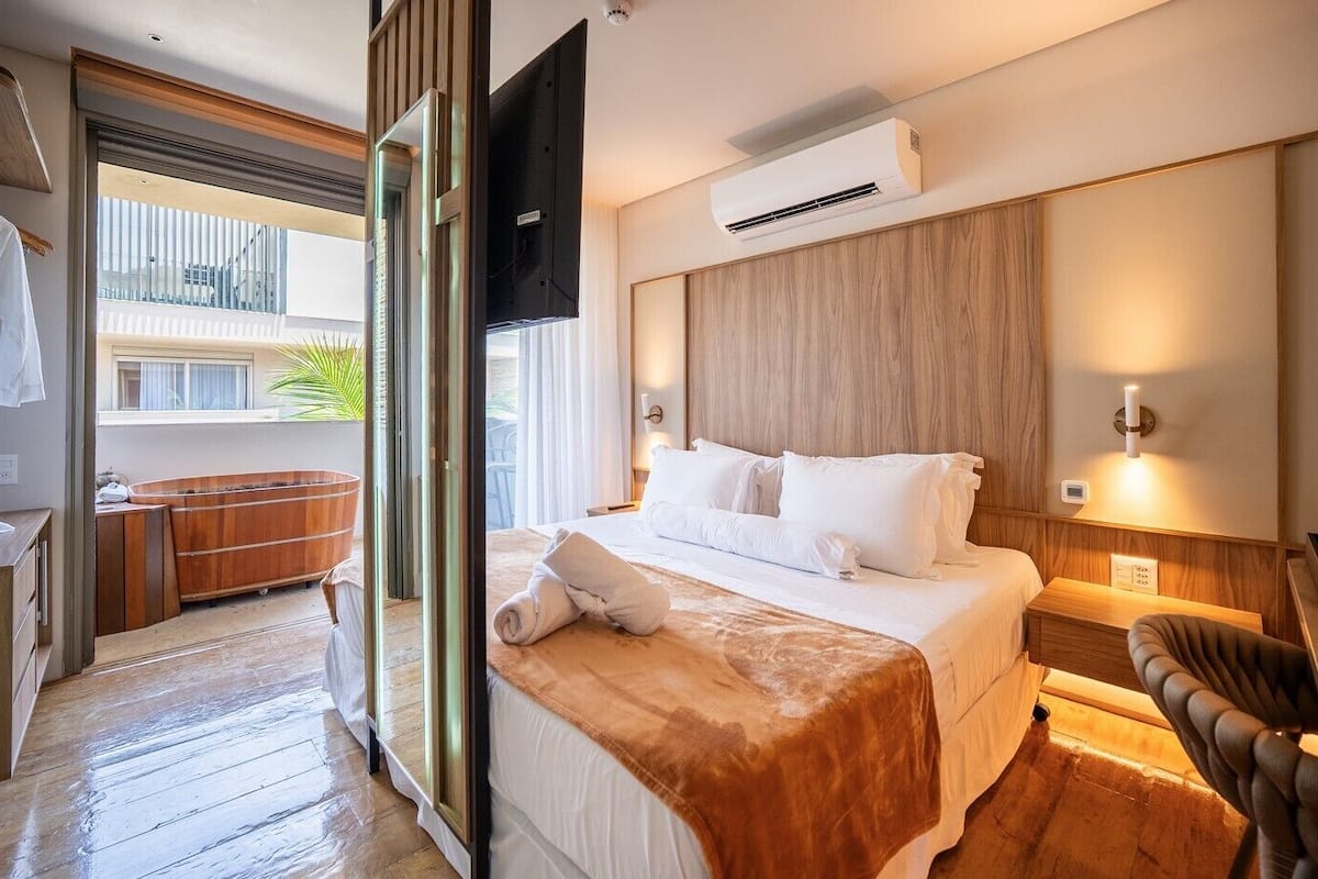 Una habitación de hotel con una cama, cabecera de madera, unidad de aire acondicionado, mesita de noche y una silla.