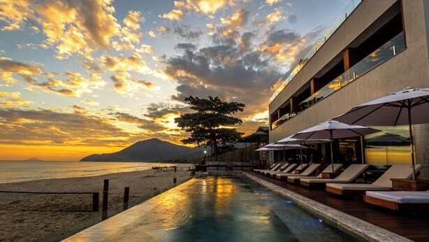 A poolside area with lounge chairs and umbrellas, overlooking a beach and ocean at sunset. - Duke Beach Hotel & SPA Maresias (São Sebastião)