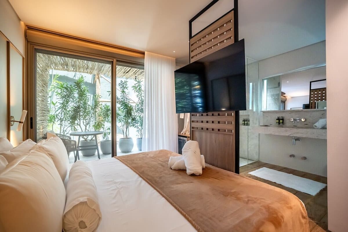 Habitación de hotel con una cama grande, televisor, baño con lavamanos y espejo, y balcón con plantas.