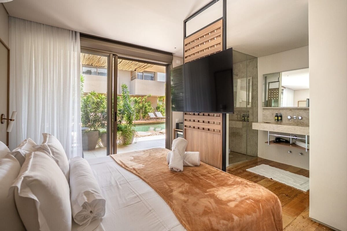Habitación de hotel moderna con cama, televisión, baño y vistas a una piscina y zonas verdes.