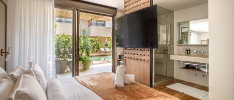 A modern hotel room with a bed, a TV, a bathroom, and a view of a pool and greenery.