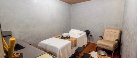 A small, compact room with a single bed, a chair, a small table, and a desk with a lamp.