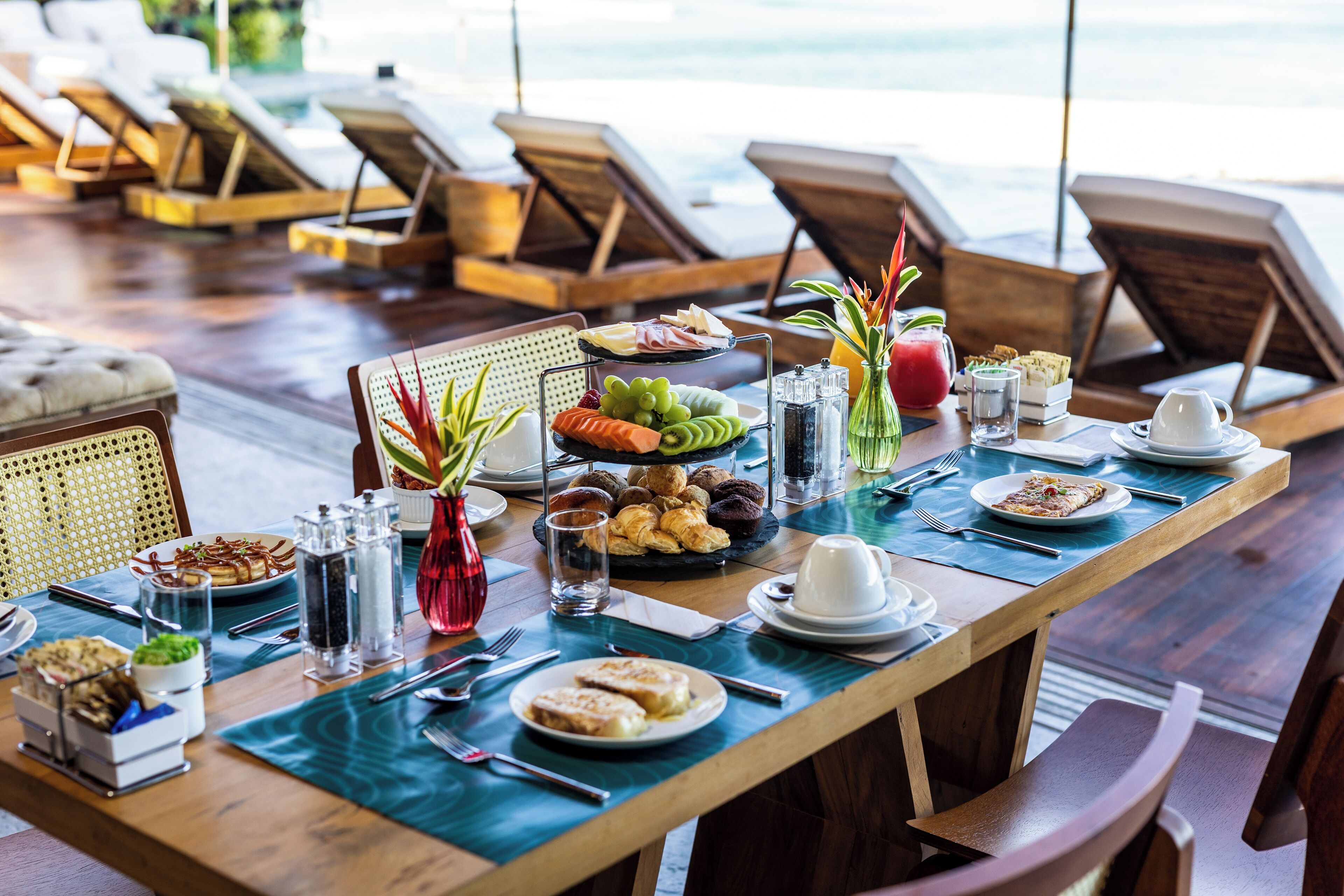 A dining table set with various breakfast items, including fruits, pastries, and beverages, in a room with wooden furniture and a view of the ocean.