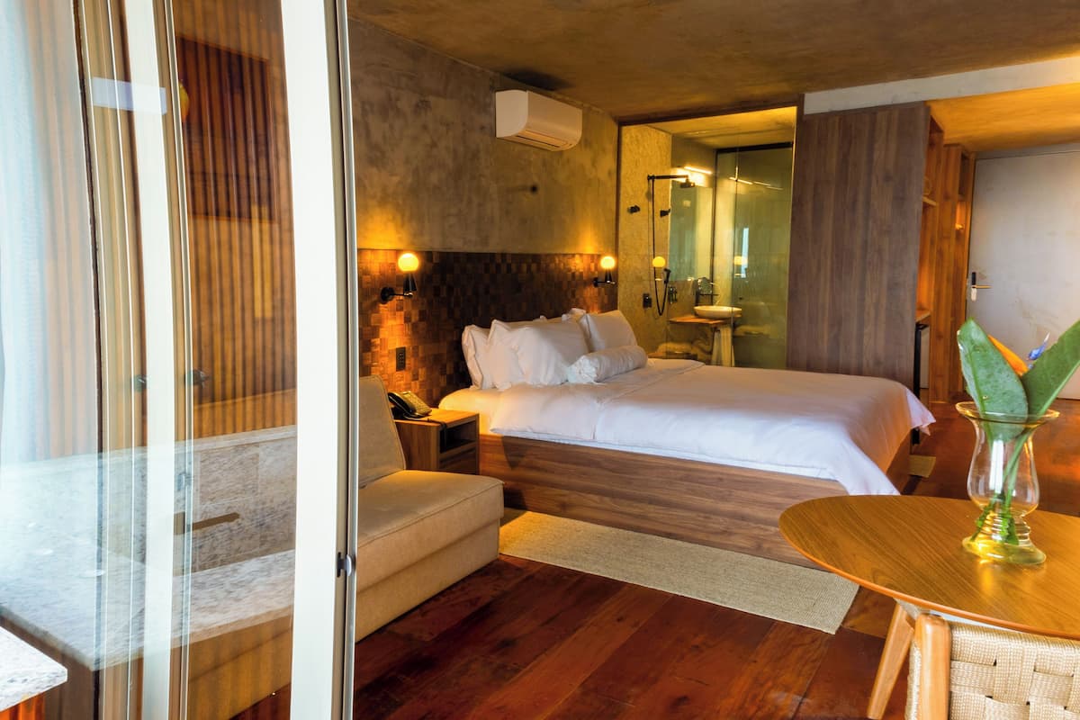 Una habitación de hotel moderna con una cama grande, mesitas de noche, una pequeña zona de estar y un baño visible a través de una puerta abierta.