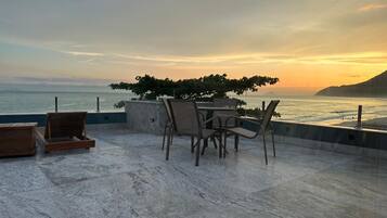 Una terraza con mesa y sillas que da al mar, con vistas a la costa y un árbol que da sombra.