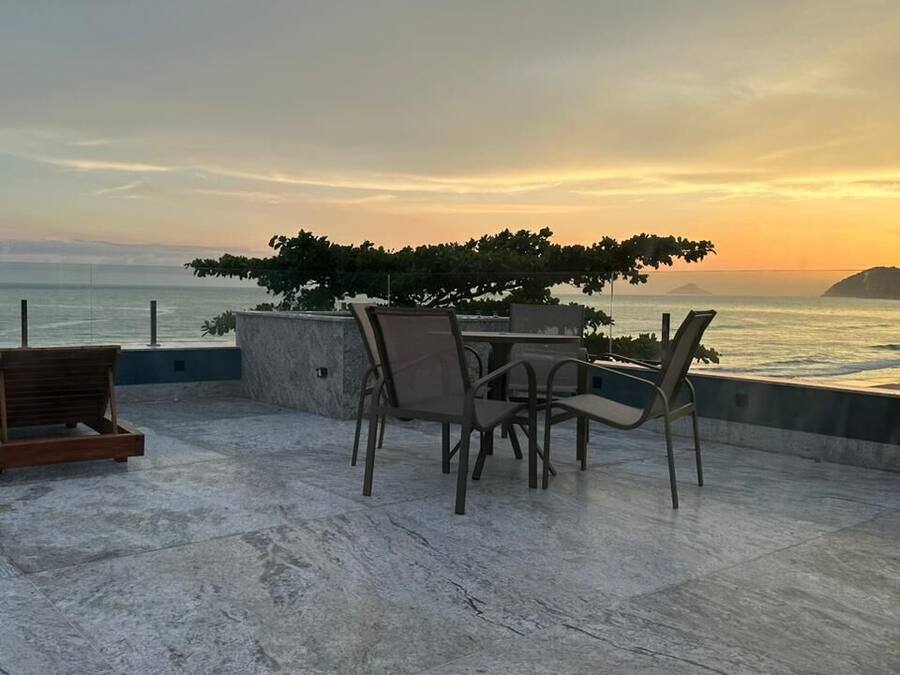 Una terraza con mesa y sillas que da al mar, con vistas a la costa y un árbol que da sombra.