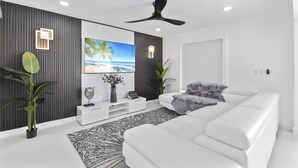 Een moderne woonkamer met een witte bank, een plafondventilator, een grote televisie en een decoratief wandpaneel.