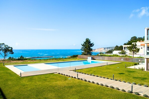 A swimming pool with a view of the ocean, surrounded by a grassy lawn and a wooden deck. - Caminho Real S - T2 Holiday Apartment with views of Nazaré beach By SCH (Nazaré)