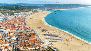 A coastal cityscape with a beach lined with rows of beach umbrellas and tents, a clear blue sea, and a dense urban area with terracotta-roofed buildings.
