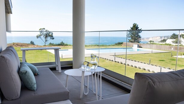 A balcony with a view of the ocean, a pool, and a seating area with cushions and a table.