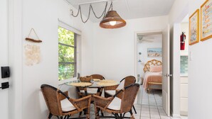 A dining area with wicker chairs and a glass table, a wicker pendant light, and a view of a bedroom with a bed and a painting. - Jetties Pointe Cottage #3 - 408   “Casey Key” (Nokomis)