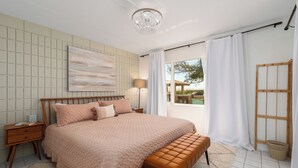 A bedroom with a large bed, a wooden bench, a nightstand, a lamp, and a view of the outdoors. - Jetties Pointe Cottage #3 - 408   “Casey Key” (Nokomis)