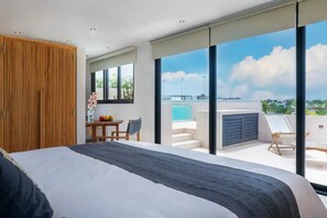 Une chambre moderne avec un grand lit, une armoire en bois, une petite table avec des fruits et une vue sur un balcon donnant sur une piscine.