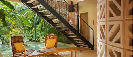 A modern interior with a staircase, two wicker chairs with yellow cushions, and a pool area visible through the glass.