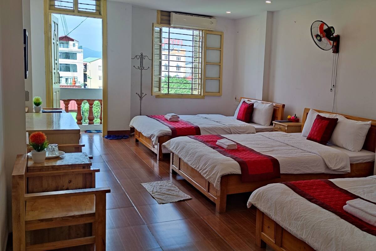 A hotel room with three beds, a wooden desk, and a window with a view of buildings.