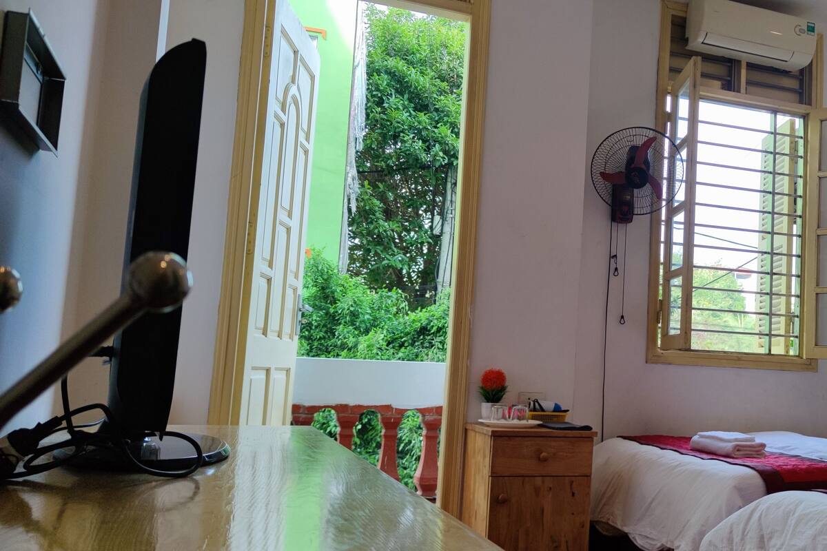 A hotel room with two beds, a desk with a computer, a window with a view of greenery, and an air conditioning unit.