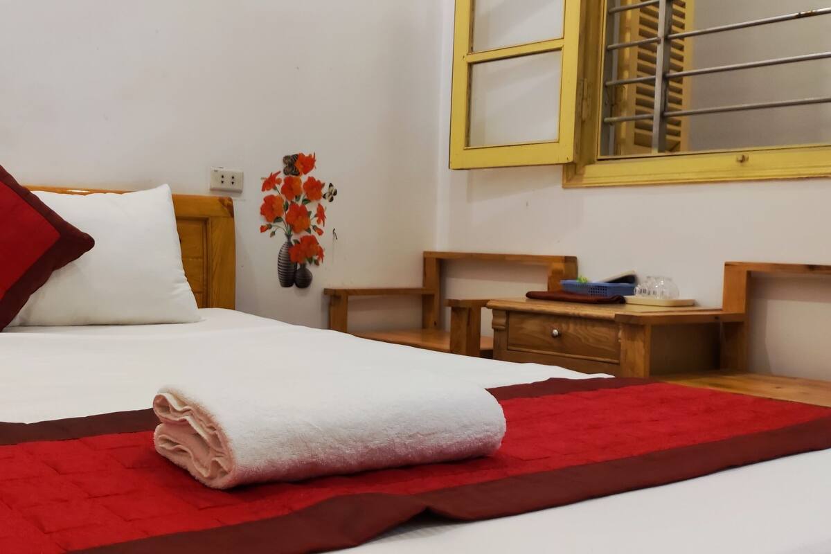 A neatly made bed with white and red bedding, a red throw blanket, and a wooden nightstand with a lamp and a vase of flowers.